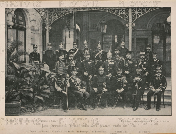 Group portrait of foreign officers, Les Officiers Étrangers aux Maneuvers de 1895, page, photomechanical print, inscription: '22 Japon. 19 France. 15 Suisse. 21 Suede. 10 Portugal. 11 Roumania. 7 Etats Unis. 20 France., 6 T... 4 Belgique', inscription: 'Extrait de la Photo-Gazette.', inscription: 'Phototype pris sur plaques Perron, à Màcon.', photographer: Henry Dufey, cliché maker: anonymous, 1895, paper, autotypie, height 115 mm x width 179 mm, Print on third unnumbered page after page 80., anonymous historical persons depicted in a group, in a group-portrait, A sepia-toned photograph shows a large group of uniformed men posed on steps and chairs beneath an ornate metal awning in front of a stone building with arched doorways and windows; many men wear peaked caps or helmets and military jackets adorned with medals, sashes, and braided cords, and several hold swords or canes; potted plants with broad leaves occupy the foreground at one side, and decorative ironwork frames the top of ...