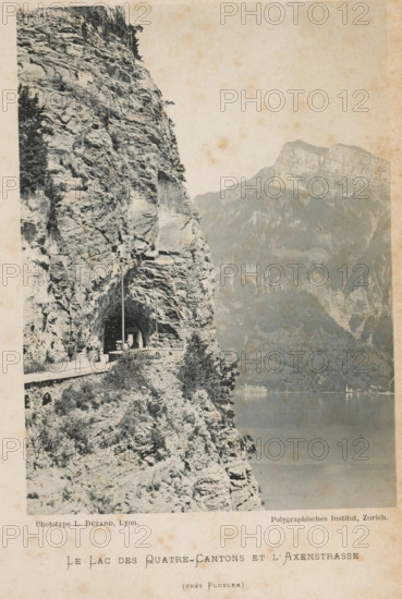 Le Lac des Quatre-Cantons et l'Axenstrasse, View of the Axenstrasse and Lake Lucerne, page, photomechanical print, maker: L. Détard, maker: Polygraphisches Institut AG, c. 1892 - in or before 1897, L. Détard, Polygraphisches Institut AG, paper, collotype, photoprint, height 163 mm x width 119 mm, lake, mountains, Lake Lucerne, Axenstrasse, A monochrome photograph shows a steep rocky cliff face with rugged strata and sparse vegetation along a narrow road carved into the rock, the road passing through a short tunnel in the cliff where a lamp post and a small figure are visible; below the cliff edge the terrain drops toward a calm lake whose surface reflects muted tones, and opposite the lake a distant mountainside with layered rock faces rises under a pale sky.