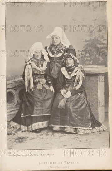Costumes de Bavière (Basse-Franconie), Portrait of three unknown women in Bavarian costumes, page, photomechanical print, cliché maker: Meisenbach, Riffarth & Cie., Munich, c. 1892 - in or before 1897, photographer, Bavaria, paper, autotypie, height 180 mm x width 125 mm, folk costume, regional costume, anonymous historical persons depicted in a group, in a group-portrait - BB - woman, Three women are posed together wearing long dresses with full skirts and layered aprons, high collars, and ornate necklaces; each has a head covering with one tall woman standing behind two seated women, the standing woman wearing a pointed white headdress that drapes down behind her and a layered scarf around her neck, the seated women wearing different shaped headpieces with ribbons and decorative bands, one seated woman resting her hand on the other's shoulder while the other clasps her hands in her lap; the dresses show patterned trims and pleated underskirts at the hems, and the backdrop includes a ...