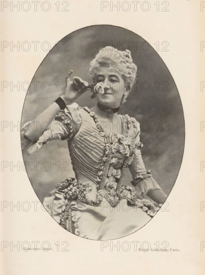 Portrait of an unknown woman, page, photomechanical print, monogram: 'AG ph.', inscription: 'Phot. Corresp.', inscription: 'Concours Suter', maker: Roger Galichon, cliché maker: unknown, c. 1894 - in or before 1899, paper, autotypie, height 136 mm x width 104 mm, historical persons not known by name - BB - woman, cut flowers, nosegay, bunch of flowers, A sepia-toned oval photograph shows a woman in a decorative, ruffled dress with layered fabric and floral trim, seated and holding a small flower near her face with one hand; she has styled, curly hair adorned with small flowers, a choker around her neck, and a faint smile, while the background within the oval is plain and softly shaded.