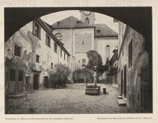 Fraumünsteramt, Courtyard of the Fraumünster Monastery in Zurich, page, photomechanical print, maker: l'Ecole professionelle de photographie, cliché maker: Meisenbach, Riffarth & Cie., Munich, c. 1895 - in or before 1900, l'Ecole professionelle de photographie, Meisenbach, cliché maker, Bavaria, paper, autotypie, height 109 mm x width 152 mm, courtyard, cloisters, monastery, Zürich (city), A cobblestone courtyard framed by a dark arched opening shows aged buildings with textured, weathered facades and shuttered windows lining both sides, a central stone fountain with a tall ornate column and rectangular basin sits in the open space, a large building with a steep roof and small dormer windows rises at the far end behind a leafy tree, narrow doorways and small steps lead into the buildings, and the scene is rendered in a monochrome, grainy photographic style.