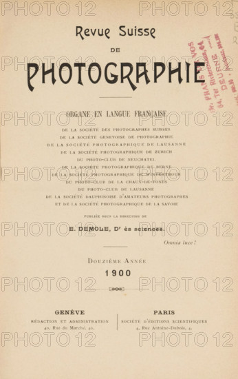 Revue suisse de photographie, Société genevoise de photographie ... et al., book, date: 'Nov 1994', annotation: '... van deveren ...', number: '288', collector's mark: 'Dr. Frans de vos, 64 tot Rivierenlaan, 64, Deurne, ...', editor (person who revises): Eugène Demole, 1900, Eugène Demole, editor (person who revises), textile materials, paper, linen (material), transparent paper, printing, autotype, collotype, height 234 mm x width 172 mm x thickness 29 mm, The image shows a vertical page with a cream background and printed elements arranged from top to bottom, including a large bold decorative word near the top, a horizontal thin line, several lines of smaller uppercase text forming centered blocks, a stamped diagonal mark in a darker color on the right side, a small ornamental divider, and two separated blocks of text near the lower portion with a vertical dividing line between them, all set against a plain background with even margins.