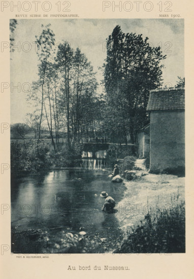 Au bord du Nusseau, View of a river with children playing, page, photomechanical print, photographer: Gottfried Wolfsgruber, cliché maker: anonymous, c. 1897 - in or before 1902, paper, collotype, photoprint, height 155 mm x width 115 mm, river, child, A vertical black-and-white photograph showing a calm river bordered by a grassy, rocky bank on the right and dense foliage and tall, thin trees on the left, with a small bridge visible in the midground and reflections of trees in the water, a building with a tiled roof occupying the right edge, and two people wearing hats seated near the water’s edge on the bank.