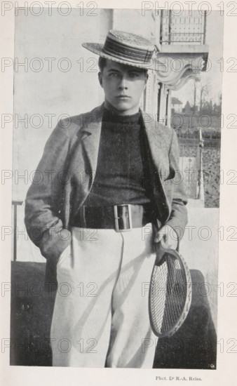 Portrait of an unknown man with a tennis racket, page, photomechanical print, monogram: 'C. Cie', photographer: Rodolphe Archibald Reiss, cliché maker: Corbaz & Cie, c. 1897 - in or before 1902, paper, autotypie, height 178 mm x width 103 mm, historical persons, tennis, lawn-tennis, A black-and-white portrait of a young person standing outdoors wearing a straw boater hat tilted at an angle, a dark high-neck sweater, a light jacket draped over the shoulders, light trousers held by a wide belt with a rectangular buckle, and one hand in a pocket while the other holds a round tennis racket with visible strings; an ornate building facade and a railing appear in the background.