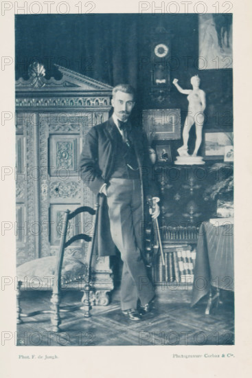 Portrait of an unknown man, page, photomechanical print, monogram: 'C. Cie', photographer: Francis de Jongh, cliché maker: Corbaz & Cie, c. 1897 - in or before 1902, paper, autotypie, height 167 mm x width 110 mm, historical persons, interior of the house, A man with a mustache and beard stands leaning against an ornate wooden chair, dressed in a dark jacket, waistcoat, trousers, and polished shoes, one hand in a pocket and the other resting on a cane; behind him is a carved wooden cabinet with intricate paneling and a tall clock mounted on the wall, a draped cloth covering a table topped with a small statue of a nude female figure on a pedestal and several framed pictures, while a stack of books sits on a lower shelf and patterned flooring is visible beneath the furniture.