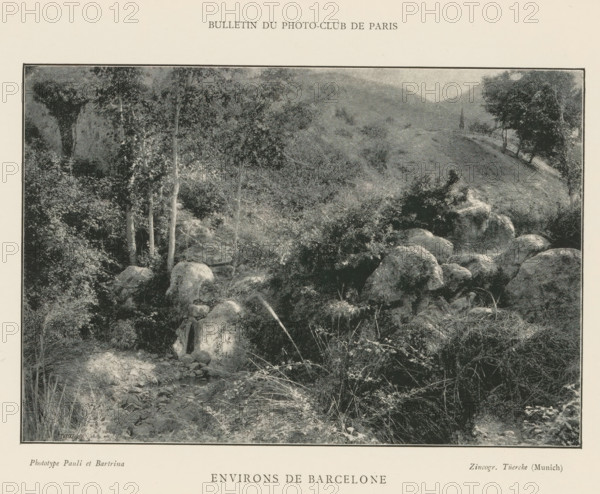 Rocky landscape near Barcelona, Environs de Barcelone, page, photomechanical print, maker: Pauli et Bartrina, cliché maker: Tüerke, Munich, c. 1890 - in or before 1895, Bavaria, paper, autotypie, height 106 mm x width 156 mm, landscapes, rock-formations, Catalonia, A monochrome landscape photograph framed by a rectangular border shows a rocky foreground with large rounded boulders and scattered low vegetation, a shallow stream or dry creek bed winding among stones, tall grasses and small shrubs interspersed, slender tree trunks rising near the left side with leafy branches, denser foliage and undergrowth filling much of the middle ground, a sloping hillside covered in low vegetation extending toward the background, and more trees silhouetted along the distant ridge under a softly mottled sky.