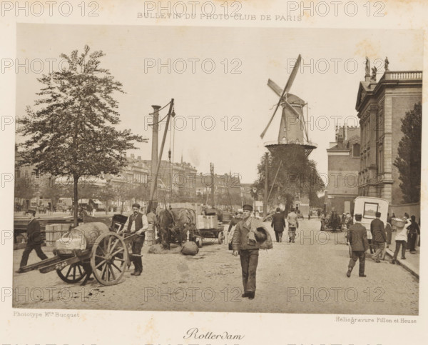 Rotterdam, View of a street in Rotterdam, with a mill in the background, page, photomechanical print, inscription: 'Bulletin du Photo-Club de Paris', photographer: Maurice Bucquet, cliché maker: Fillon & Heuse, Paris, c. 1890 - in or before 1895, paper, photoaquatint, height 120 mm x width 170 mm, street, windmill, A street scene showing a cobblestone road with people walking and standing, some carrying items and one man walking toward the camera holding a bundled object over his shoulder, horse-drawn carts and wooden wagons loaded with barrels and crates, a large tree on the left, a windmill with visible sails near the center-right, a tall building with classical architectural details and statues on its roof on the right, lamp posts and poles with hanging equipment, rows of multi-story buildings in the background, and various street activity with figures in period clothing.