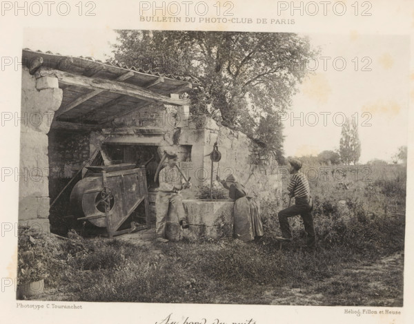 At the well, Two men and a woman around a well, page, photomechanical print, inscription: 'Bulletin du Photo-Club de Paris', maker: C. Touranchet, cliché maker: Fillon & Heuse, Paris, printer (person): Charles Wittmann, c. 1890 - in or before 1895, printer (person), paper, autotype, height 120 mm x width 170 mm, adult woman, adult man, well, A grayscale photograph shows an outdoor scene with a stone structure on the left featuring a tiled overhang and wooden beams, and a large leafy tree rising behind it; in front of the structure there is an old mechanical device with a large wheel and a trough or basin set into a low stone wall; three people stand by the basin, one wearing a cap and light clothing leaning against the wall holding a curved implement, another bending over the basin with a skirt and headscarf, and a third in a striped top and cap standing on the grass with one foot on a low step holding a pole; the foreground is grassy with some low vegetation and a potted plant at the ...