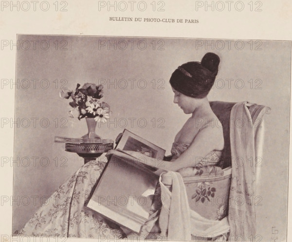 Liseuse, Portrait of an unknown woman reading, page, photomechanical print, monogram: 'AG ph.', maker: Constant Puyo, cliché maker: unknown, printer (person): Draeger & Lesieur, Paris, c. 1890 - in or before 1895, printer (person), paper, autotypie, height 119 mm x width 162 mm, historical persons not known by name - BB - woman, reading, A sepia-toned photograph of a seated woman in profile, wearing her hair in a high bun and a strapless patterned dress, holding and looking at an open large book on her lap; she sits in an upholstered armchair draped with a long fabric, and beside her is a small round table with a vase of mixed flowers on top.