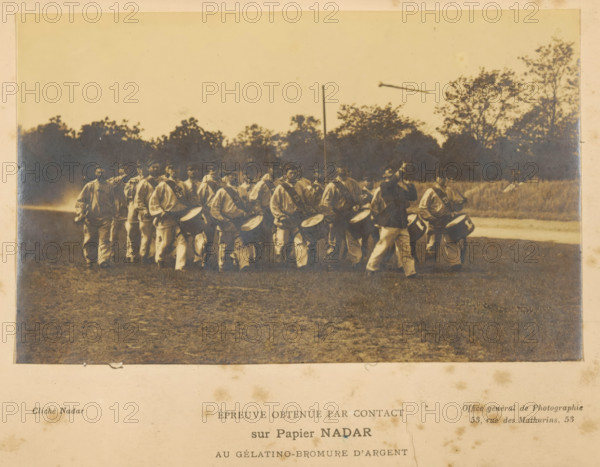 View of a group of unknown men with, among other things, drums, photograph, page, inscription: 'Office general de Photographie, 53, Rue de Mathurins, 53', inscription: 'Cliché Nadar', inscription: 'Épreuve obtenue par cohtact, sur Papier NADAR, au gélatino-bromure d'argent', photographer: Félix Nadar, photographer: Paul Nadar, c. 1883 - in or before 1888, Félix Nadar, Paul Nadar, paper, baryta paper, gelatin silver print, developing gelatin silver print, height 77 mm x width 124 mm, military band, A sepia-toned photograph shows a group of people standing and marching on a grassy field with trees in the background, many of them holding or wearing drums and drum straps, dressed in light-colored clothing with darker accents, and one figure in a darker outfit positioned slightly forward and turning with an arm raised, while a vertical pole rises behind them and a horizontal object appears in the sky above the treeline.