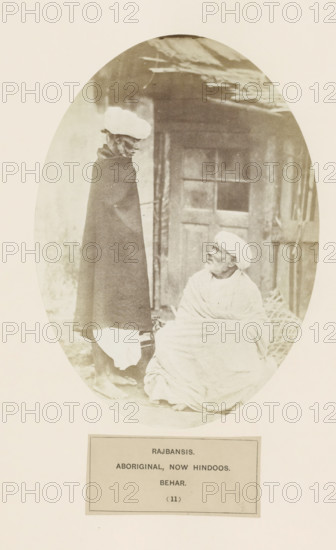 Two men with turbans from the Rajbanshi community of Bihar in eastern India, photograph, page, number: '(11)', inscription: 'RAJBANSIS., ABORIGINAL, NOW HINDOOS., BEHAR.', Bihar, c. 1858 - in or before 1868, photographer, North India, paper, albumen print, height 195 mm x width 140 mm, human races, peoples, nationalities, An oval-framed sepia photograph showing two men in traditional clothing in front of a wooden doorway; one man stands wearing a dark long cloak and a light turban, facing slightly toward the seated man, and the seated man is wrapped in a light shawl with a light turban, looking up at the standing figure; a woven basket rests near the seated man and the doorway shows a paneled door and rough wooden beams above.