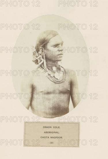 Portrait of an unknown man from the Kurukh community of Chota Nagpur in eastern India, photograph, page, The Kurukh community is also known as Oraon., number: '(15)', inscription: 'ORAON COLE., ABORIGINAL., CHOTA NAGPOOR.', photographer: Benjamin Simpson (possibly), Chota Nagpur, c. 1858 - in or before 1868, Jharkhand, paper, albumen print, height 159 mm x width 120 mm, human races, peoples, nationalities, An oval-framed sepia portrait of a bare-chested person with smooth, dark hair pulled back and adorned with decorative elements at the side, wearing multiple layered necklaces and pendants, looking to the right with a neutral expression against a plain light background, with a rectangular label positioned below the portrait.
