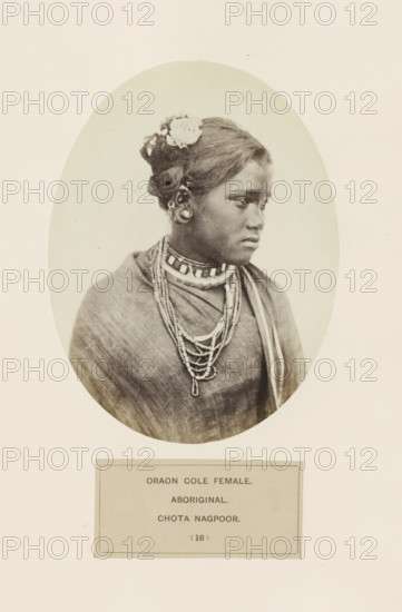 Portrait of an unknown woman from the Kurukh community of Chota Nagpur in eastern India, photograph, page, The Kurukh community is also known as Oraon., inscription: 'ORAON COLE FEMALE., ABORIGINAL., CHOTA NAGPOOR.', number: '(16)', photographer: Benjamin Simpson (possibly), Chota Nagpur, c. 1858 - in or before 1868, Jharkhand, paper, albumen print, height 159 mm x width 120 mm, human races, peoples, nationalities, A sepia-toned oval portrait of a young woman shown from the chest up, turned slightly to her left with a neutral expression, wearing a draped garment and multiple layered beaded necklaces, her hair styled up with a flower tucked near the top and decorative ear ornaments visible, set against a plain light background and mounted on a larger sheet of paper.