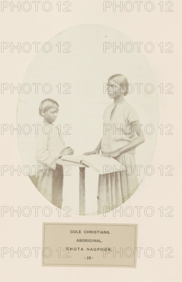Praying boy and a girl with a book behind a desk from Chota Nagpur in eastern India, photograph, page, The missionary's work resulted in the conversion of the tribal population to Christianity., number: '(19)', inscription: 'COLE CHRISTIANS., ABORIGINAL., CHOTA NAGPOOR.', photographer: Benjamin Simpson (possibly), Chota Nagpur, c. 1858 - in or before 1868, Benjamin Simpson, photographer, Jharkhand, paper, albumen print, height 175 mm x width 130 mm, one person praying, human races, peoples, nationalities, An oval-framed sepia photograph shows two young people standing at a small round table with an open book; the person on the left is shorter, looking downward with hands held together near the book, wearing a long-sleeved light top and a skirt, with short hair; the person on the right is taller, standing upright with one hand resting on the book and the other near the table, wearing a short-sleeved light top and a long skirt, with hair pulled back and a composed facial expression; the ...