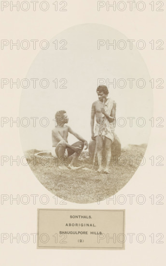 Two unknown men from the Santhal tribe in eastern India, photograph, page, inscription: 'SONTHALS., ABORIGINAL., BHAUGULPORE HILLS.', number: '(2)', Bhagalpur, c. 1858 - in or before 1868, photographer, North India, Bihar, paper, albumen print, height 175 mm x width 130 mm, human races, peoples, nationalities, Jharkhand, Rajmahal hills, An oval-framed sepia photograph shows two barefoot men on grassy ground, one man seated with knees bent and arms resting on them, wearing a short cloth around his waist and looking up toward the standing man, while the standing man wears a draped cloth over one shoulder and around his waist, holds a long wooden staff vertically, and stands beside a large rounded earthen vessel partially visible behind him; the background inside the oval is plain and light, and the image is mounted on a pale rectangular sheet.