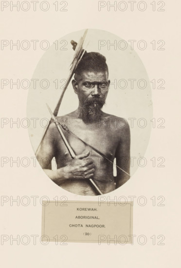 Portait of an unknown man with a bow and arrow from the Korwa tribe of Chota Nagpur in eastern India, photograph, page, number: '(20)', inscription: 'KOREWAH., ABORIGINAL., CHOTA NAGPOOR.', photographer: Benjamin Simpson, Chota Nagpur, c. 1858 - in or before 1868, Jharkhand, paper, albumen print, height 160 mm x width 121 mm, archer's weapons: bow and arrow, human races, peoples, nationalities, An oval black-and-white portrait of a bare-chested man with a narrow face and a beard, his hair tied up, holding a long spear diagonally across his chest and a shorter pointed implement in his hand, with a neutral light background and a rectangular label area below the oval.