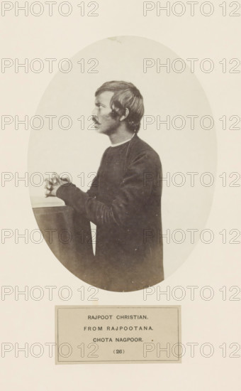 A praying Christian man from Rajput community in a kneeling position in Chota Nagpur, eastern India, photograph, page, inscription: 'RAJPOOT CHRISTIAN., FROM RAJPOOTANA., CHOTA NAGPOOR.', number: '(26)', photographer: Benjamin Simpson (possibly), Chota Nagpur, c. 1858 - in or before 1868, Jharkhand, paper, albumen print, height 175 mm x width 129 mm, one person praying, human races, peoples, nationalities, A sepia-toned oval photograph shows a young man in profile facing left, with short dark hair and a mustache, wearing a dark long-sleeved garment with a lighter collar visible at the neck; he is posed with his hands clasped and resting on a flat surface, and the oval image is set against a pale rectangular background with a separate rectangular label area below the portrait.