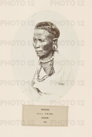 Portrait of an unknown man from the Miri tribe of Assam, photograph, page, The Miri tribe is also known as Mising., inscription: 'MEEREE., HILL TRIBE., ASSAM.', number: '(30)', photographer: attributed to Benjamin Simpson, Assam, c. 1858 - in or before 1868, Attributed to, paper, albumen print, height 175 mm x width 130 mm, human races, peoples, nationalities, An oval-framed black-and-white portrait of an older person shown from the chest upward, facing slightly left with a neutral expression and visible facial wrinkles, a short straight fringe haircut, large circular ear ornaments, multiple layered necklaces including a strand of shell-like beads, a draped light-colored cloth over the shoulders, and one hand holding the cloth near the chest; the portrait sits on a pale background with a rectangular label area below.