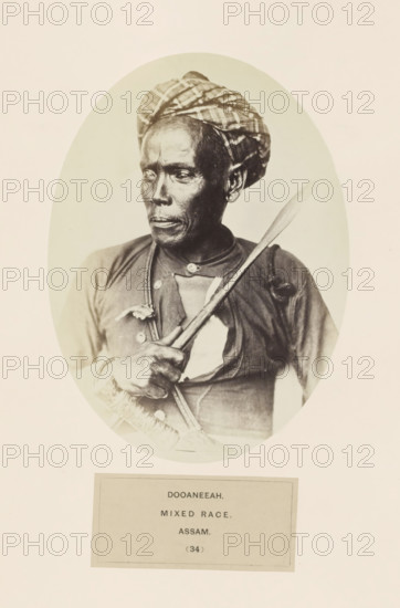 Portrait of an unknown man with a spear from the Dowania tribe of Assam, photograph, page, number: '(34)', inscription: 'DOOANEEAH., MIXED RACE., ASSAM.', photographer: Benjamin Simpson (possibly), Assam, c. 1858 - in or before 1868, paper, albumen print, height 175 mm x width 129 mm, head-gear: turban, human races, peoples, nationalities, casting weapons: spear, A sepia-toned oval portrait of a person with dark skin wearing a patterned headwrap and a buttoned garment, shown from the chest up and turned slightly to the viewer's left, holding a long narrow object diagonally across the chest with one hand gripping its handle, the face showing a neutral expression with visible facial lines and an earring in one ear, the image set against a pale background with a rectangular label area below the oval.