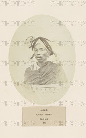 Portrait of an unknown man from the Kuki tribe of Assam, photograph, page, number: '(39)', inscription: 'KOOKIE., ROBBER TRIBES., CACHAR.', photographer: Benjamin Simpson (possibly), Cachar, c. 1858 - in or before 1868, Assam, paper, albumen print, height 175 mm x width 129 mm, human races, peoples, nationalities, A faded oval portrait of a person wearing a patterned headwrap with fabric tassels and a decorative floral adornment, visible earrings, and a draped garment; the person is seated behind a carved wooden or woven object with a scalloped edge, and the portrait is mounted on a pale background with a rectangular label panel below.