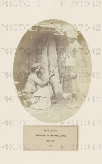 Two unknown men with a rifle from the Malik clan of Bihar in eastern India, photograph, page, inscription: 'MULLICKS., SOONEE MAHOMEDANS., BEHAR.', number: '(5)', Bihar, c. 1858 - in or before 1868, photographer, North India, paper, albumen print, height 159 mm x width 120 mm, human races, peoples, nationalities, An oval sepia photograph shows two men in front of a rustic wall and thatched roof; one man is seated on the ground with a wrapped lower garment and a cloth around his head, holding a long curved object toward the other man, while the standing man wears a long wrapped skirtlike garment, a cloth turban, and a wide belt around his waist, looking down at the seated man; the ground appears bare and a bundle of sticks leans against the wall behind them.