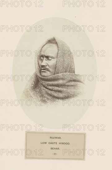 Portrait of an unknown man from the Rajwar community of Bihar in eastern India, photograph, page, number: '(7)', inscription: 'RUJWAR., LOW CASTE HINDOO., BEHAR.', Bihar, c. 1858 - in or before 1868, photographer, North India, paper, albumen print, height 160 mm x width 120 mm, human races, peoples, nationalities, A sepia-toned oval portrait of a person wrapped in a textured shawl that covers the head and shoulders, showing a close-up three-quarter profile with a serious facial expression, short hair visible at the forehead, prominent cheekbones, a mustache, and deep-set eyes looking toward the left, against a plain light background and surrounded by a wide blank border.
