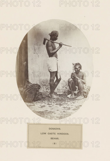 Two unknown men with tools from the Dusadh community of Bihar in eastern India, photograph, page, The Dusadh community is also known as Paswan., inscription: 'DOSADHS., LOW CASTE HINDOOS., BEHAR.', number: '(9)', Bihar, c. 1858 - in or before 1868, photographer, North India, paper, albumen print, height 158 mm x width 121 mm, human races, peoples, nationalities, A sepia-toned oval photograph shows two barefoot men against a plain wall. One man stands with a cloth wrapped around his waist and a headscarf, holding a long-handled tool resting on his shoulder and looking downward. The other man sits on the ground with a cloth around his lower body, holding a small object vertically in both hands and looking at it. There are cloth bundles and scattered debris on the ground near their feet.