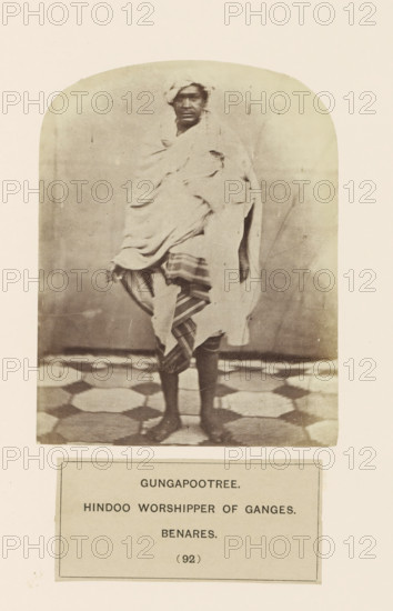 Portrait of an unknown man from the Gangaputra Brahmin community of Varanasi, Uttar Pradesh, India, photograph, page, inscription: 'GUNGAPOOTREE., HINDOO WORSHIPPER OF GANGES., BENARES.', number: '(92)', Varanasi, c. 1858 - in or before 1868, photographer, North India, paper, albumen print, height 122 mm x width 95 mm, Hinduism, Buddhism, Jainism, human races, peoples, nationalities, A full-length sepia photograph of a person standing on a patterned floor, wrapped in layered light-colored drapery with a striped cloth visible around the lower body, a cloth wrapped around the head, bare legs and feet showing, and a plain backdrop with rounded top framing the image.