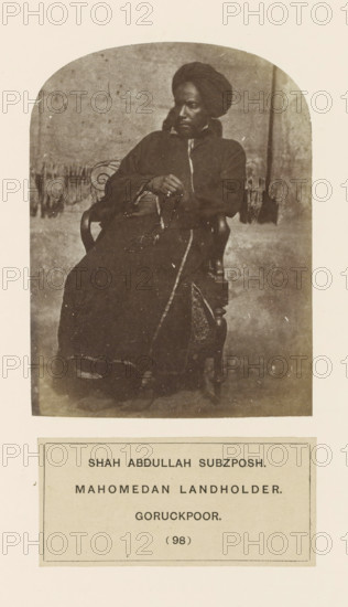 Portrait of Shah Abdullah Subzposh from Gorakhpur, Uttar Pradesh, India, photograph, page, number: '(98)', inscription: 'SHAH ABDULLAH SUBZPOSH., MAHOMEDAN LANDHOLDER., GORUCKPOOR.', Gorakhpur, c. 1858 - in or before 1868, photographer, North India, paper, albumen print, height 117 mm x width 88 mm, human races, peoples, nationalities, A sepia-toned photograph shows a seated person wearing a dark, full-length garment and a wrapped head covering, leaning slightly back in a wooden chair with ornate armrests; the person's hands rest in their lap holding a string of beads and a thin stick, and a patterned fabric panel is visible on the chair beside them, while a plain backdrop and a fringed textile behind the chair are seen in the upper portion of the image and a blank margin surrounds the photograph.
