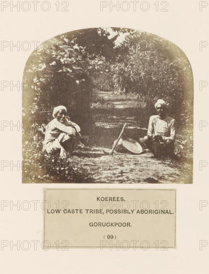 Two unknown men on a path with agricultural tools from the Kori community of Gorakhpur, Uttar Pradesh, India, photograph, page, inscription: 'KOEREES., LOW CASTE TRIBE, POSSIBLY ABORIGINAL., GORUCKPOOR.', number: '(99)', Gorakhpur, c. 1858 - in or before 1868, photographer, North India, paper, albumen print, height 98 mm x width 108 mm, road, path, human races, peoples, nationalities, agricultural implements, A sepia-toned photograph framed by a rounded arch shows two seated individuals on a dirt path surrounded by dense foliage; one person sits cross-legged on the left wearing light clothing and a headwrap with arms resting on their knees, while the other on the right also wears a headwrap and light clothing and sits with hands in their lap; between them on the ground lies a long wooden stick leaning against a shallow round metal pan, and the background features leafy trees and undergrowth fading into shadow.