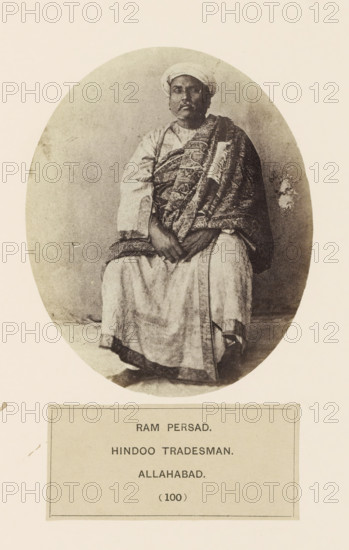 Portrait of a tradesman Ram Persad from Prayagraj, Uttar Pradesh, India, photograph, page, Prayagraj is also known as Allahabad., inscription: 'RAM PERSAD., HINDOO TRADESMAN., ALLAHABAD.', number: '(100)', Allahabad, c. 1858 - in or before 1868, photographer, North India, paper, albumen print, height 119 mm x width 97 mm, head-gear: turban, human races, peoples, nationalities, persons engaged in trade and commerce, A sepia-toned oval photograph shows a seated man wearing a turban and traditional layered garments, including a patterned draped cloth over one shoulder and a lighter robe underneath; his hands rest together in his lap and his feet are visible beneath the long robe, with a plain background surrounding him and a rectangular label area positioned below the oval.