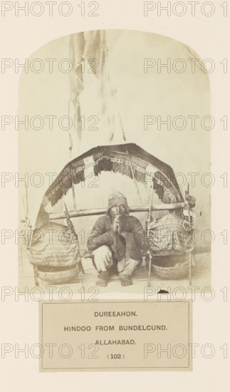 Portrait of an unknown man from Bundelkhand region who carries water from the Ganges in a bamboo scaffolding in Prayagraj, Uttar Pradesh, India, photograph, page, Prayagraj is also known as Allahabad., inscription: 'DUREEAHON., HINDOO FROM BUNDELCUND., ALLAHABAD.', number: '(102)', Allahabad, c. 1858 - in or before 1868, photographer, North India, paper, albumen print, height 147 mm x width 107 mm, human races, peoples, nationalities, A monochrome photograph shows a seated man squatting with his forearms resting on his knees and his hands held near his face, wearing a cap and layered clothing with loose trousers and bare feet; he sits beneath a woven canopy with fringed edges supported by a horizontal pole, flanked on each side by large woven baskets or bundles hanging from the pole and resting on low supports, with additional vertical poles and coiled rope visible on the right, and a plain light background filling the upper portion of the image.