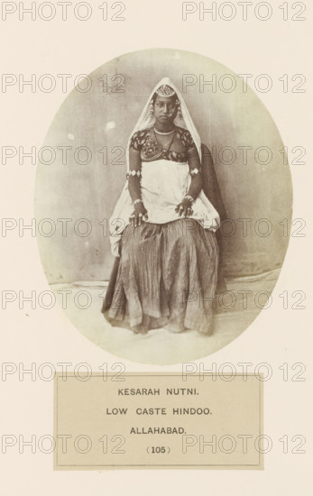 Portrait of Kesarah Nutni, a woman from Prayagraj, Uttar Pradesh, India, photograph, page, Praragraj is also known as Allahabad., inscription: 'KESARAH NUTNI., LOW CASTE HINDOO., ALLAHABAD.', number: '(105)', Allahabad, c. 1858 - in or before 1868, photographer, North India, paper, albumen print, height 129 mm x width 105 mm, human races, peoples, nationalities, A sepia oval photograph of a seated woman wearing a veil over her head and shoulders, a patterned short-sleeved blouse, and a draped cloth around her torso, with a long pleated skirt covering her legs; she has multiple pieces of jewelry including necklaces, bracelets, and rings, and decorative ornaments on her forehead and hair, with her hands resting on her knees and a neutral facial expression, set against a plain backdrop and mounted on a page with a rectangular label beneath the oval.