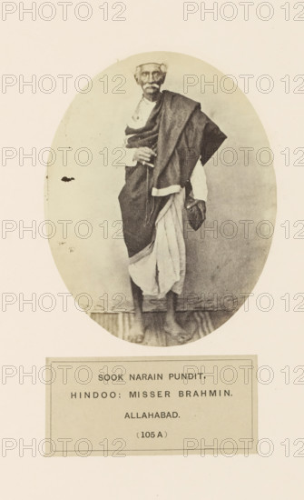Portrait of Sook Narain, a high priest from Prayagraj, Uttar Pradesh, India, photograph, page, Prayagraj is also known as Allahabad., number: '(105 A )', inscription: 'SOOK NARAIN PUNDIT., HINDOO: MISSER BRAHMIN., ALLAHABAD.', Allahabad, c. 1858 - in or before 1868, photographer, North India, paper, albumen print, height 120 mm x width 95 mm, human races, peoples, nationalities, priest, Hinduism, Buddhism, Jainism, An oval sepia photograph shows an older man standing on a narrow wooden floor, wearing a wrapped garment draped over one shoulder and a light cloth around his lower body, with a cloth cap on his head and a shawl folded across his arm, his feet are bare and his hands are held near his midsection, and the oval image is set against a light rectangular background with a bordered panel beneath it.