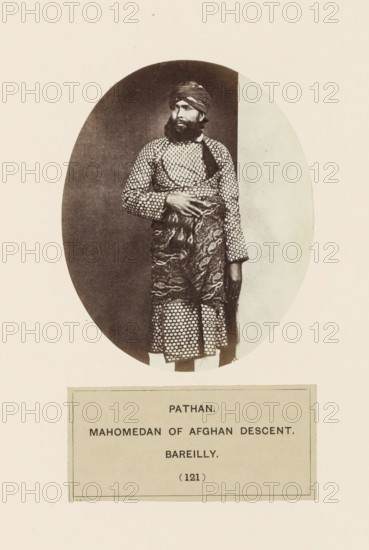 Portrait of an unknown man from the Pathan community of Bareilly, Uttar Pradesh, India, photograph, page, number: '(121)', inscription: 'PATHAN., MAHOMEDAN OF AFGHAN DESCENT., BAREILLY.', Bareilly, c. 1858 - in or before 1868, photographer, North India, paper, albumen print, height 107 mm x width 88 mm, human races, peoples, nationalities, A sepia-toned oval photograph shows a bearded man standing against a plain backdrop, wearing a patterned long tunic and a patterned wrap around his waist, with a turban on his head; his left arm hangs at his side while his right hand rests on the wrapped cloth at his waist, and the image is mounted on a light-colored page with a rectangular label beneath the photograph.