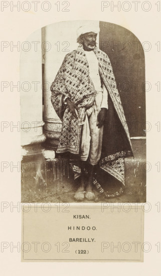 Portrait of an unknown man from the Kisan community of Bareilly, Uttar Pradesh, India, photograph, page, inscription: 'KISAN., HINDOO., BAREILLY.', number: '(122)', Bareilly, c. 1858 - in or before 1868, photographer, North India, paper, albumen print, height 127 mm x width 88 mm, human races, peoples, nationalities, A sepia photograph shows a standing man wearing a patterned shawl draped over his shoulders and wrapped around his body, a light turban on his head, a light long-sleeved shirt, and loose light trousers; he has a beard and mustache, his hands rest near his hips, and he stands barefoot on a step in front of a column and a dark doorway, with the edge of the patterned shawl trailing to the ground.