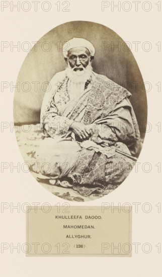 Portrait of Khulleefa Daood from Aligarh, Uttar Pradesh, India, photograph, page, inscription: 'KHULLEEFA DAOOD., MAHOMEDAN., ALLYGHUR.', number: '(126)', Aligarh, c. 1858 - in or before 1868, photographer, North India, paper, albumen print, height 147 mm x width 109 mm, human races, peoples, nationalities, An oval sepia photograph of an elderly man seated cross-legged on a patterned cloth, wearing a turban and layered robes with intricate patterns, a long white beard, and hands resting in his lap, framed by a pale border with a rectangular panel below.