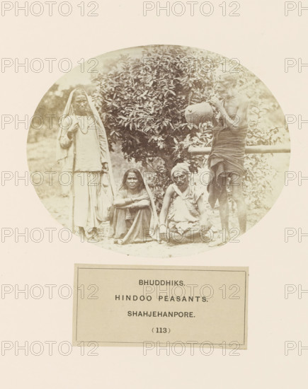 Four unknown Bhuddhiks from Shahjahanpur in front of a bush, Bhuddhiks. Hindu peasants. Shahjehanpore, photograph, page, number: '(113)', Shahjahanpur, c. 1858 - in or before 1868, photographer, North India, Uttar Pradesh, paper, albumen print, height 104 mm x width 129 mm, human races, peoples, nationalities, A faded circular photograph shows four people positioned in front of a leafy bush, with two individuals seated on the ground and two standing. On the left a person stands wearing a long light garment with a cloth draped over the head and shoulders, holding a staff and a bag. In the center foreground two seated figures face the camera; one has a cloth covering the head and rests arms on folded knees, the other wears a wrapped head covering and a draped upper garment with hands resting on the ground. On the right a thin standing figure wears a wrapped lower cloth and a head covering while holding a rounded pot near the face. The background includes foliage and a horizontal wooden r...