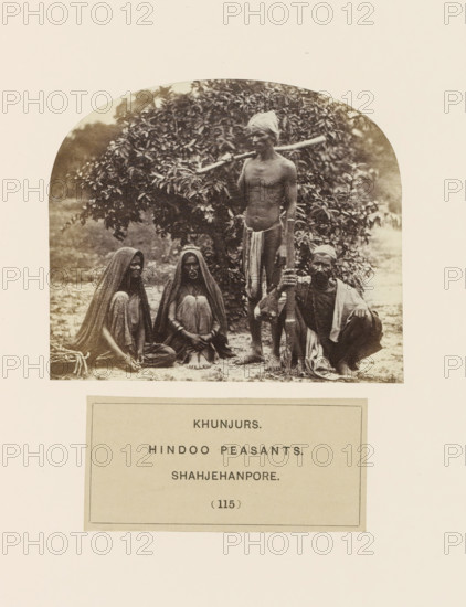 Four people from the Kanjar tribe of Shahjahanpur, Uttar Pradesh, India, photograph, page, number: '(115)', inscription, `: 'KHUNJURS., HINDOO PEASANTS., SHAHJEHANPORE', Shahjahanpur, c. 1858 - in or before 1868, photographer, North India, paper, albumen print, height 92 mm x width 107 mm, farmers, human races, peoples, nationalities, A sepia photograph framed by a rounded top shows four people posed outdoors in front of dense foliage; two seated women on the left wear long draped garments covering their heads and bodies and sit with knees drawn up, one of them displaying a large circular ornament on the face, a standing man in the center is bare-chested with a cloth wrapped around his head and a long piece of cloth tied around his waist while he rests a long pole across his shoulders, and a crouching man on the right wears a headwrap and loose garments and holds a tall staff upright with one hand.