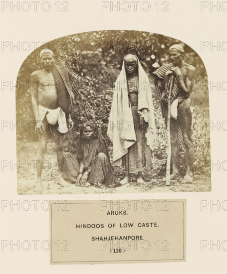 Four people from the Arakh community of Shahjahanpur, Uttar Pradesh, India, photograph, page, inscription: 'ARUKS., HINDOOS OF LOW CASTE., SHAHJEHANPORE.', number: '(116)', Shahjahanpur, c. 1858 - in or before 1868, photographer, North India, paper, albumen print, height 101 mm x width 120 mm, human races, peoples, nationalities, A sepia-toned photograph shows four people posed outdoors against a backdrop of foliage; two are standing on either side, one stands in the center with a long draped cloth over the head and shoulders, and one sits on the ground in front with hands resting on the knees. The standing figures on the sides wear cloth wraps around their waists and have additional fabric draped over one shoulder, one holding a long wooden staff. The seated person wears patterned lower garments and a head covering, with bare feet visible. The central standing figure’s draped cloth falls to the feet and partially covers the body. The ground beneath them appears uneven and covered with...