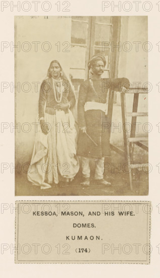 Portrait of a couple in front of a house in Kumaon, Uttarakhand, India, photograph, page, number: '(174)', inscription: 'KESSOA, MASON, AND HIS WIFE., DOMES., KUMAON.', Kumaon, c. 1859 - in or before 1869, photographer, paper, albumen print, height 113 mm x width 85 mm, couple of lovers, human races, peoples, nationalities, A faded sepia photograph shows a man and a woman standing side by side in front of a building with a paneled window; the woman on the left wears layered clothing including a long patterned skirt, a draped outer cloth, long sleeves, necklaces, and a head covering, while the man on the right wears a long tunic with a sash at the waist, a turban, leggings, and holds a stick in one hand with his other arm resting on a wooden ladder positioned beside him.