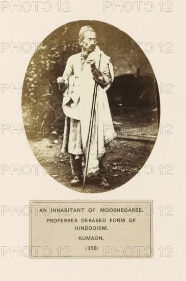 Portrait of an unknown man from the Munsyari region of Kumaon, Uttarakhand, India, photograph, page, inscription: 'AN INHABITANT OF MOOSHEGARHEE., PROFESSES DEBASED FORM OF, HINDOOISM., KUMAON.', number: '(176)', Munsyari, c. 1859 - in or before 1869, photographer, paper, albumen print, height 118 mm x width 95 mm, Hinduism, Buddhism, Jainism, human races, peoples, nationalities, A sepia-toned oval photograph shows an older man standing outdoors on a dirt surface, wearing layered traditional clothing including a long garment and a draped shawl, with a small cap on his head; he holds a long staff with one hand and a small object in the other, and his face shows a mustache and beard while he looks slightly to the side, with blurred foliage and dark background behind him.