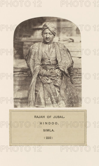 Portrait of the raja (king) of Jubbal near Shimla, Himachal Pradesh, India, photograph, page, number: '(210)', inscription: 'RAJAH OF JUBAL., HINDOO., SIMLA.', Shimla, c. 1859 - in or before 1869, photographer, paper, albumen print, height 100 mm x width 76 mm, ruler, sovereign, human races, peoples, nationalities, A sepia-toned portrait of a seated person wearing a patterned robe and a turban, hands clasped in their lap, shown inside a rounded-arch frame above a rectangular label panel.