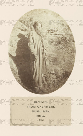 Portrait of an unknown merchant from Kashmir in Shimla, Himachal Pradesh, India, photograph, page, number: '(215)', inscription: 'CASHMIRI., FROM CASHMERE., MUSSULMAN., SIMLA.', Shimla, c. 1859 - in or before 1869, photographer, paper, albumen print, height 120 mm x width 95 mm, merchant, salesman, head-gear: turban, human races, peoples, nationalities, A sepia-toned oval photograph shows a bearded man standing against a rocky backdrop. He wears a long robe and a turban, and his hands rest near a walking staff. The rock surface behind him casts a shadow, and grasses or small plants are visible on the uneven ground. The image is mounted on a pale page with a rectangular label area below the oval.