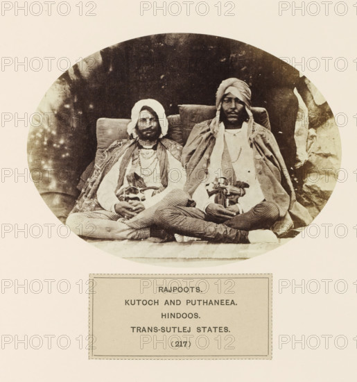 Portrait of two unknown men from the Rajput community of Kangra, Himachal Pradesh, India, photograph, page, number: '(217)', inscription: 'RAJPOOTS., KUTOCH AND PUTHANEEA., HINDOOS., TRANS-SUTLEJ STATES.', Kangra, c. 1859 - in or before 1869, photographer, paper, albumen print, height 109 mm x width 147 mm, head-gear: turban, human races, peoples, nationalities, A circular sepia photograph shows two seated men wearing layered clothing and headwraps, each seated against cushioned backs and holding curved-hilted swords across their laps, with patterned fabrics draped over their shoulders and striped trousers visible where their legs cross.