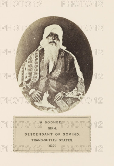 Portrait of an unknown man from the Sodhi community of Kangra, Himachal Pradesh, India, photograph, page, number: '(219)', inscription: 'A SODHEE., SIKH., DESCENDANT OF GOVIND., TRANS-SUTLEJ STATES.', c. 1859 - in or before 1869, photographer, paper, albumen print, height 108 mm x width 86 mm, head-gear: turban, human races, peoples, nationalities, An oval sepia photograph shows an older man seated, wearing a light headwrap and a long white beard, with a patterned shawl draped over his shoulders and a darker coat beneath; his hands rest on his lap and the background inside the oval is dark while the surrounding page is pale.