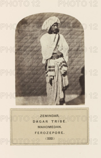 Portrait of an unknown landowner from Firozpur, Punjab, India, photograph, page, number: '(222)', inscription: 'ZEMINDAR., DAGAR TRIBE., MAHOMEDAN., FEROZEPORE.', Firozpur, c. 1859 - in or before 1869, photographer, paper, albumen print, height 100 mm x width 75 mm, head-gear: turban, human races, peoples, nationalities, A monochrome photograph of a standing man wearing layered loose garments and a large wrapped turban, his arms crossed at waist level, with a long draped cloth hanging from his waist and patterned bands on the lower fabric, set against a plain textured backdrop and framed by a rounded arch, above a bordered rectangular label containing printed lines.