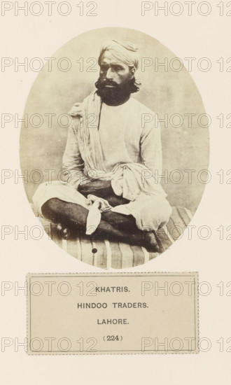 Portrait of an unknown trader from the Khatri community of Lahore, Punjab, Pakistan, photograph, page, inscription: 'KHATRIS., HINDOO TRADERS., LAHORE.', number: '(224)', Lahore, c. 1859 - in or before 1869, photographer, paper, albumen print, height 120 mm x width 95 mm, head-gear: turban, human races, peoples, nationalities, persons engaged in trade and commerce, A sepia-toned oval photograph of a seated man wearing a turban and loose light-colored clothing, with a long beard and mustache, sitting cross-legged on a striped cushion or mat, one arm resting on his knee and fabric draped over his shoulder and lap, set against a plain background and mounted on a pale page with a rectangular bordered label beneath the oval.