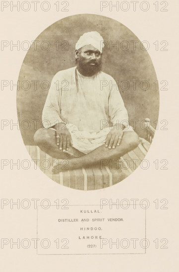 Portrait of a distiller and seller of liquor (kullal) from Lahore, Punjab, Pakistan, photograph, page, number: '(227)', inscription: 'KULLAL., DISTILLER AND SPIRIT VENDOR., HINDOO., LAHORE.', Lahore, c. 1862 - in or before 1872, photographer, paper, albumen print, height 129 mm x width 104 mm, distilled spirits, human races, peoples, nationalities, A sepia-toned oval photograph shows a seated man wearing a light-colored turban and loose long-sleeved garment, sitting cross-legged on a striped woven cot; his hands rest on his knees, he has a dark beard and mustache, and the plain backdrop is textured and neutral in tone.