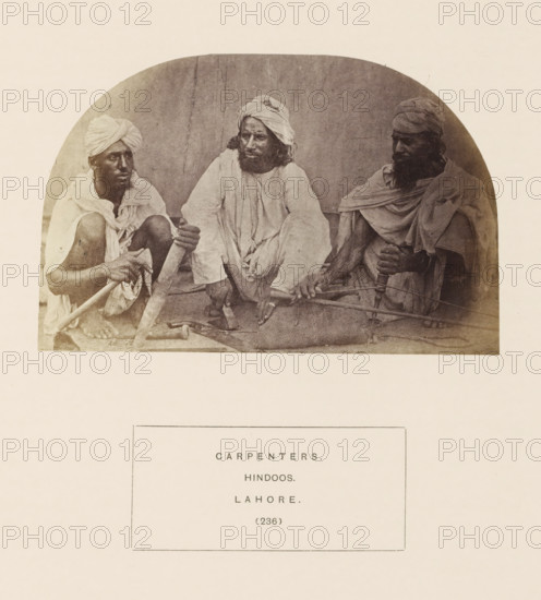 Portrait of three unknown carpenters with tools from Lahore, Punjab, Pakistan, photograph, page, inscription: 'CARPENTERS., HINDOOS., LAHORE.', number: '(236)', Lahore, c. 1862 - in or before 1872, photographer, paper, albumen print, height 89 mm x width 136 mm, carpenter, human races, peoples, nationalities, Three men sit close together on a mat against a plain backdrop, each wearing draped cloth garments and turbans; the man on the left is squatting with bare feet visible and holds a long wooden pole across his lap, the central man sits with legs bent and hands resting on the mat, looking slightly toward the left while his clothing hangs in loose folds, and the man on the right sits with a long beard and grasps a rounded wooden handle attached to a curved tool resting across his knees.